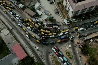 A road junction seen from directly above: yellow minibuses queued nose to tail along a curve, cars and lorries filling the lanes beside them, low roofs and stalls packed along both sides.