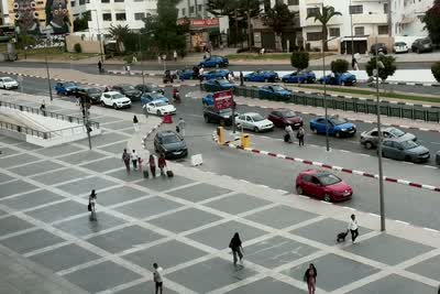 A wide paved plaza with people walking across it, and beyond a low barrier a road lined with parked cars and blue taxis.