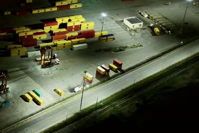 Stacks of yellow and red shipping containers lit by floodlights at night, a straddle carrier standing over a lane beside them, and an empty road running past.