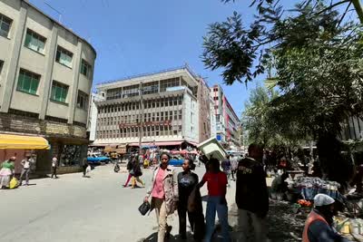 A busy shopping street: people walking past shopfronts under a yellow awning, one carrying a load on her head, mid-rise buildings and a tree behind them.